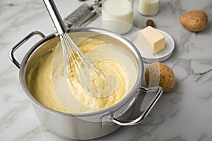 Ooking pot with mashed potatoes and ingredients as potato, milk, salt, butter, nutmeg with whisker and grinder on light marble