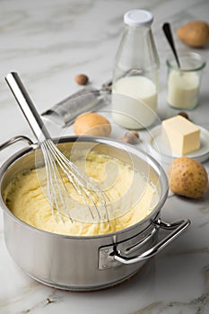 Ooking pot with mashed potatoes and ingredients as potato, milk, salt, butter, nutmeg with whisker and grinder on light marble