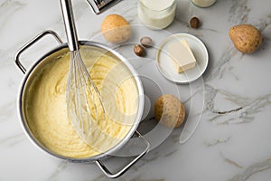 Ooking pot with mashed potatoes and ingredients as potato, milk, salt, butter, nutmeg with whisker and grinder on light marble