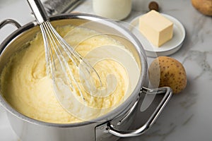 Ooking pot with mashed potatoes and ingredients as potato, milk, salt, butter, nutmeg with whisker and grinder on light marble