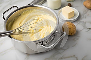 Ooking pot with mashed potatoes and ingredients as potato, milk, salt, butter, nutmeg with whisker and grinder on light marble