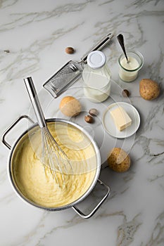 Ooking pot with mashed potatoes and ingredients as potato, milk, salt, butter, nutmeg with whisker and grinder on light marble