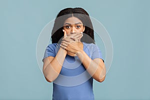Oops. African american woman covering her mouth with both hands and staring at camera against blue studio background.