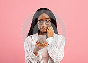 Oops. Embarassed black woman looking at mobile phone screen in confusion on pink studio background