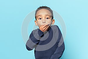 Oops. Portrait of amazed african american little boy covering mouth with hand and looking at camera, blue studio wall