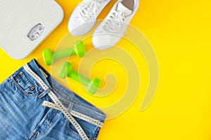 Op view photo of blue jeans with tape measure, scales, dumbbells and white sports shoes on yellow background