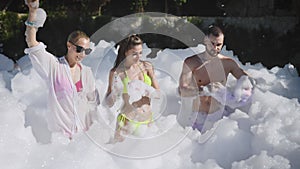 An open-air summer foam party is in full swing. Two girls in swimsuits and a young man dance to the music and have a