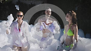 An open-air summer foam party is in full swing. Two girls in swimsuits and a young man dance to the music and have a