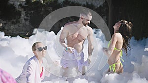 An open-air summer foam party is in full swing. Two girls in swimsuits and a young man dance to the music and have a