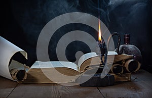 An open ancient book, a burning candle in a dark candlestick, and unfolded and rolled scrolls on a wooden table in a haze