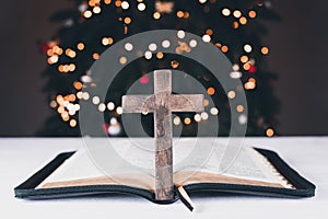 An open Bible on the table. Prayer. Wooden cross. Crucifix. On the background of the Christmas tree. Light and garlands