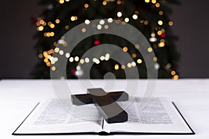 An open Bible on the table. Prayer. Wooden cross. Crucifix. Holy. On the background of the Christmas tree. Light and garlands
