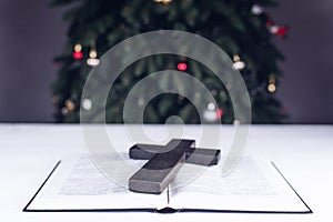 An open Bible on the table. Prayer. Wooden cross. Crucifix. Holy. On the background of the Christmas tree. Light and garlands