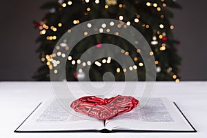 An open Bible on the table. Red heart on the book. A symbol of God\'s love. On the background of the Christmas tree.