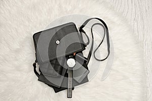 Open black bag with dropped things, notebook, mobile phone, watch, purse and lipstick. The white fur on background, top view