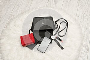 Open black bag with dropped things, notebook, mobile phone, watch, red purse and lipstick. The white fur on background, top view.