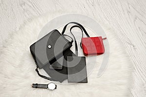 Open black bag with dropped things, notebook, mobile phone, watch and red purse. The white fur on background, top view. fashion co