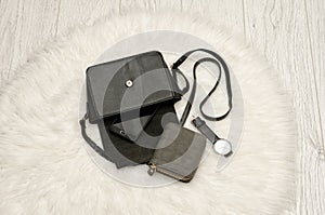 Open black bag with a spill out of her phone, a gray purse and a watch. The white fur on background, top view. fashion concept