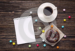 Open blank notebook, cup of coffee and a chocolate donut on a wooden table. Space for text.