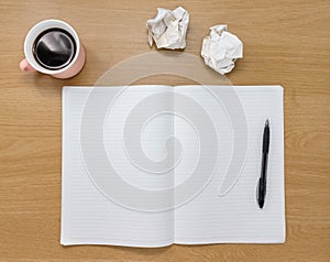 Open blank notebook, scrunched up paper and coffee cup on a wooden desk.