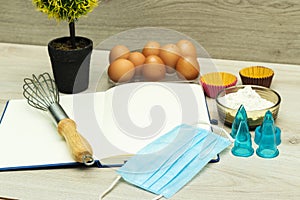 Open blank recipe book, eggs and flour, medical mask on wood table