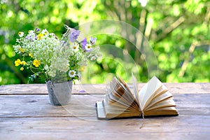 Open book in black cover, family bible, bouquet of wild flowers on wooden table in garden, blurred natural landscape in background