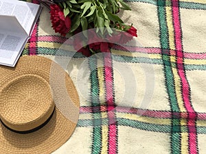 Open book, bouquet of red flowers, straw hat on the checkered white blanket. Copy space. Picnic concept