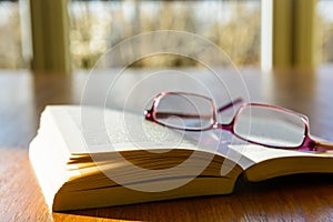 Open book and glasses on a wooden table with sunlight