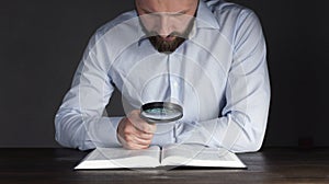 Open book Holy Bible. A man in a shirt sits at a table holding a magnifying glass looks into a book. In the dark. Businessman.