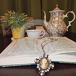 open book, necklace and medallion as a bookmark.  in the background a teapot, a tea cup, flowers and old books