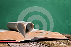 Open book and pages folded in the shape of a heart on an old wooden surface on a backdrop of green chalkboard with chalk stains,