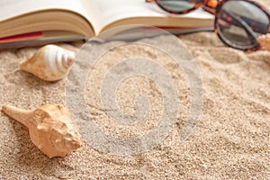 Open book on sandy beach with sea shells and sunglasses, close up lifestyle image