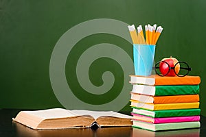 Open book with stack of books near empty green chalkboard