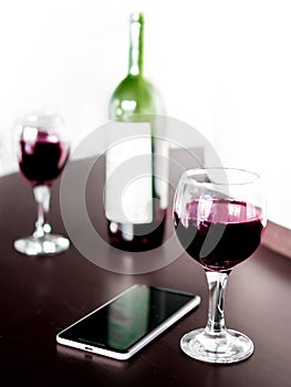 Open bottle and two glasses of red wine and a mobile phone on brown wodden table and white blurred wall background. Alcohol drink
