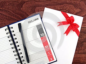 open calendar diary with date of December and January in grid and pen and envelope with red ribbon bow on desk floor
