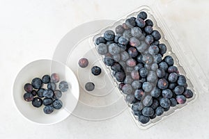 Open clamshell container of blueberries on a white granite counter, small white bowl