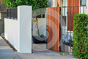 Open courtyard gate with vertical wooden pannels on frence with cement white wall and front yard trees in a neighborhood