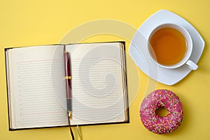 Open diary with pen, cup with tea and a sweet donut on a yellow