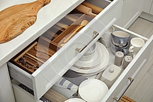 Open drawers of kitchen cabinet with different dishware, utensils and towels