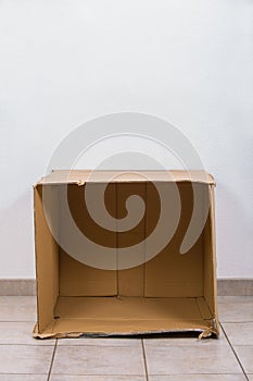 Open empty cardboard box on the floor inside a room with copy space on the wall above. Concept of improvised shelter of homeless