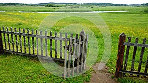 An open gate in a wooden fence and a green meadow beyond it, the path in the frame . Cloudy summer or late spring
