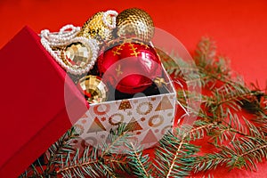 Open gift box with red and golden Christmas baubles and pearl beads on the red background. Near the spruce branch