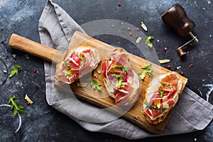 Open ham sandwiches, arugula and hard cheese, served on  wooden stand with aglass of red wine on aconcrete old dark background. Ru