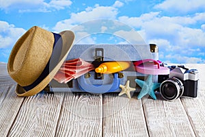 Open holiday suitcase; summer hat, towel, tanning oil, photo camera; flip flops on wooden table