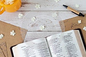 Open holy bible book with a cup of tea, old paper, pen, and white flowers on wooden table, top view