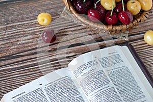 Open Holy Bible Book of Galatians with fresh red and yellow cherries in a wicker basket on a wooden table