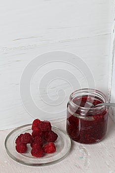 Open jar with raspberry jam. In her teaspoon. Next to a plate of fresh raspberries