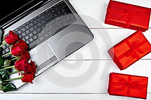 Open laptop computer, bouquet of red roses and gift boxes on white wooden desk in office, copy space. Flat lay, top view.