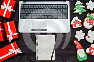 Open laptop computer, Christmas presents, blank notebook and gingerbread cookies on dark wood background, copy space.