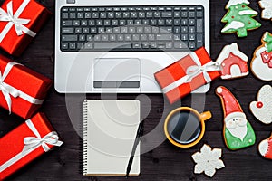 Open laptop computer keyboard, red gift boxes, blank white notebook, Christmas gingerbread cookies and cup of hot coffee.